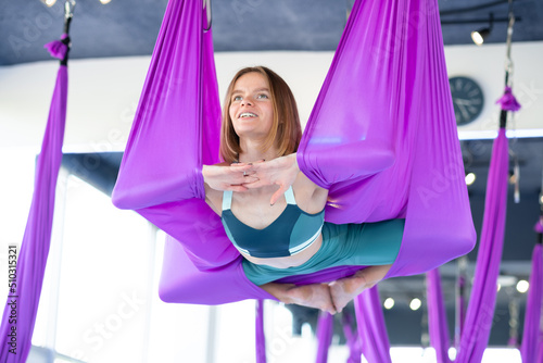 Young beautiful woman practicing aerial yoga stretching in purple hammock swing in fitness studio, healthy lifestyle and balance training, wellness and flexibility exercise