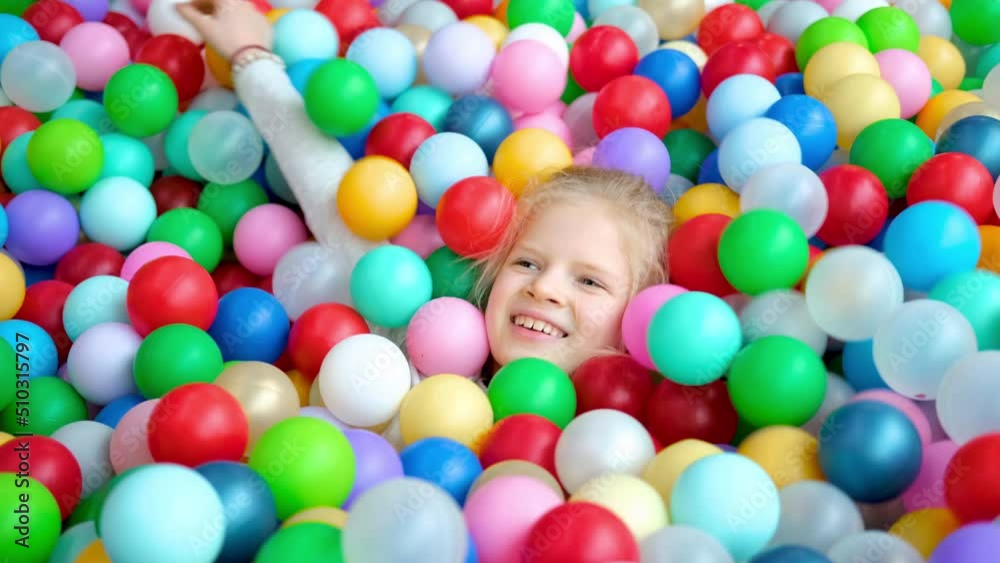 Cute blonde little girl lying on multi coloured plastic balls in big dry paddling pool in playing centre. Smiling at camera. Portrait close up. Having fun in playroom. Leisure Activity.
