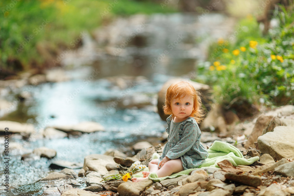 Cute little girl is sitting near the stream, space for text. Red hair baby is spending time on nature Cute little girl is sitting near the stream, space for text. Red hair baby is spending time on nature