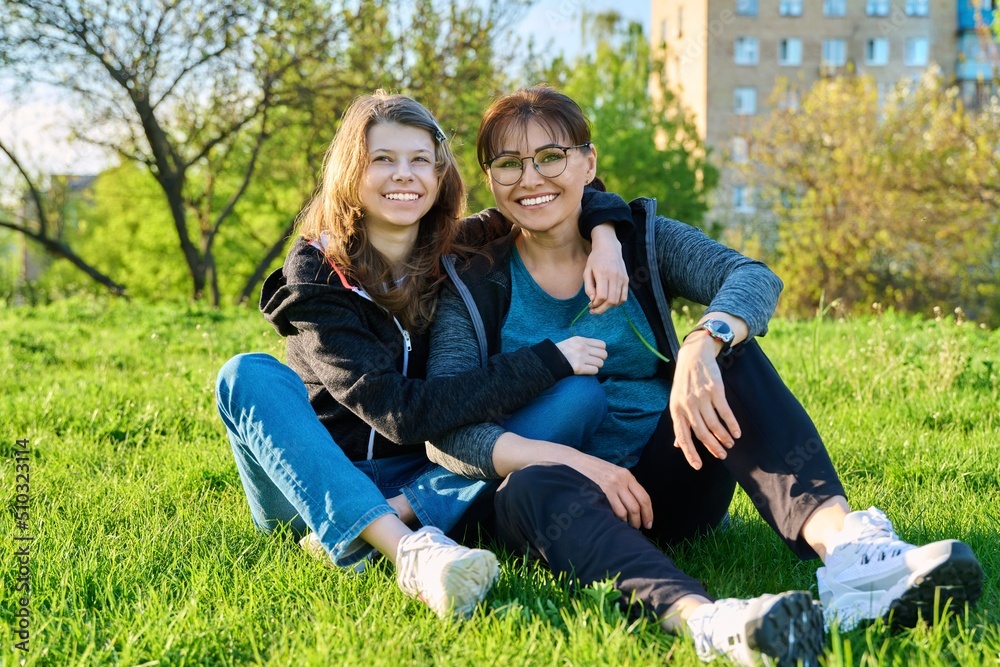 Happy mother and daughter hugging sitting on grass, looking at camera Happy mother and daughter hugging sitting on grass, looking at camera
