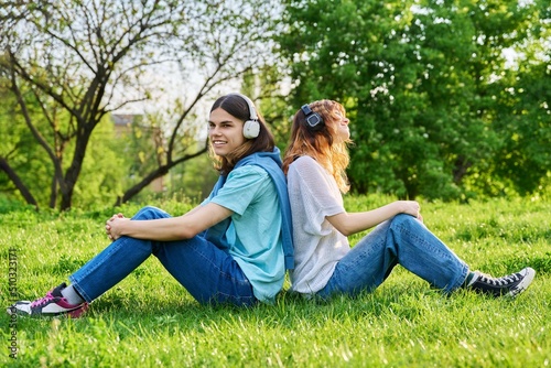 Two friends guy girl listen to music podcast, in wireless headphones, sitting on grass