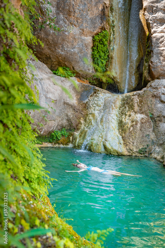 Young happy woman floating in turquoise blue crystal clear water in the river with a waterfall summer vacation relaxing body and mind.