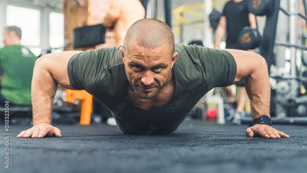 Fototapeta premium Bald caucasian tired manwearing a green t-shirt trying hard to do a push-up on gym flooring, looking directly at a camera. High quality photo