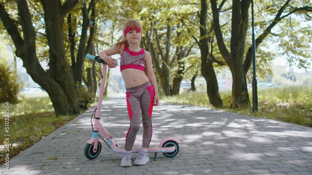 Athletic fit young girl rides on electric scooter on road in park on sunny day, outdoors. Child going standing near bike looking at camera. Kid leisure activity, sport. Modern technology for children Athletic fit young girl rides on electric scooter on road in park on sunny day, outdoors. Child going standing near bike looking at camera. Kid leisure activity, sport. Modern technology for children