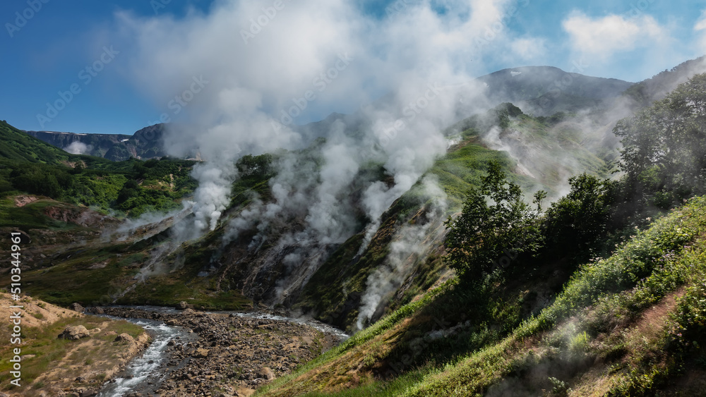 Fototapeta premium Columns of steam from hot springs rise above the slopes of the mountain. The stream flows in a hollow along a rocky bed. Blue sky. Kamchatka. Valley of Geysers