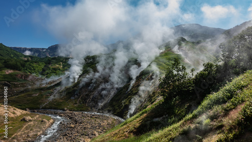 Columns of steam from hot springs rise above the slopes of the mountain. The stream flows in a hollow along a rocky bed. Blue sky. Kamchatka. Valley of Geysers