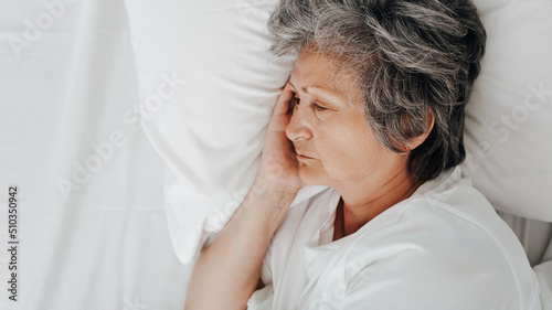 Sad senior woman lying on bed in bedroom, top view. Side view tired lonely elderly woman resting indoors and looking away, copy space. Frustration, depression, laziness, female mental health concept