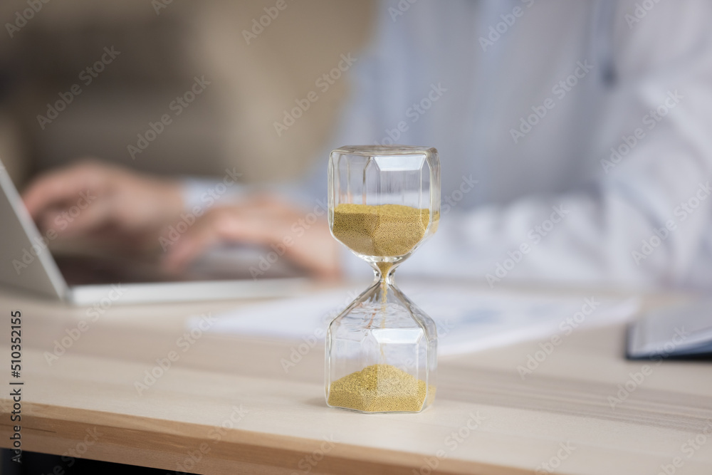 Unknown businesswoman sit at desk working on laptop, focus on hourglass showing passage of workday time. Deadline, time is money, billable hours, productive and effective work day, workflow concept
