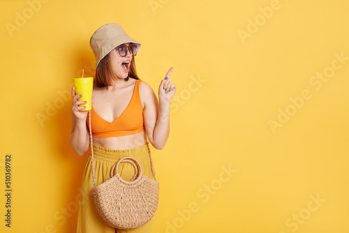 Indoor shot of astonished amazed woman in sunglasses wearing summer clothing posing isolated over yellow background, standing with fresh in hadns and pointing aside and advertising area.