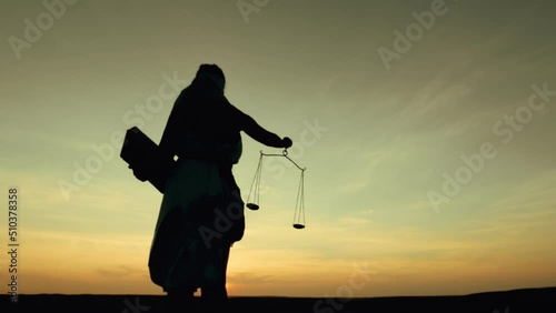 Goddess of justice stands background sunset sky and holds scale and sword. White dress of Greek goddess justice Themis flutters in wind, back view. photo