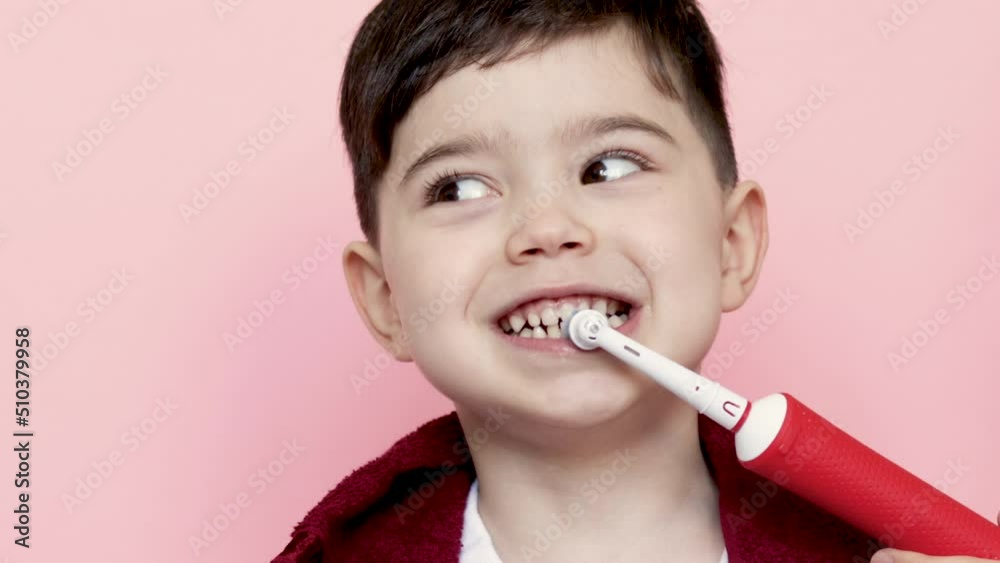 cute little boy using electric toothbrush for daily teeth cleaning.kid with dark red towel on neck.pink background,4k close up.dental care,dentist for kids concept. smiling boy, diastema,space between cute little boy using electric toothbrush for daily teeth cleaning.kid with dark red towel on neck.pink background,4k close up.dental care,dentist for kids concept. smiling boy, diastema,space between