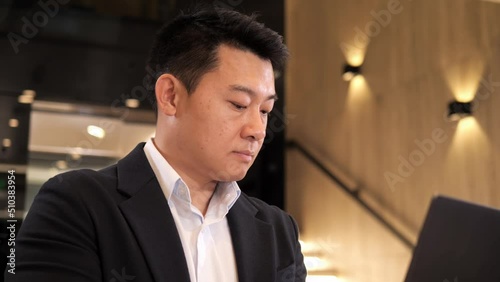 Serious asian businessman is typing message checking info using laptop in business center lobby. Portrait of focused man wearing formal black suit standing in hall with computer. An urgent matter. photo