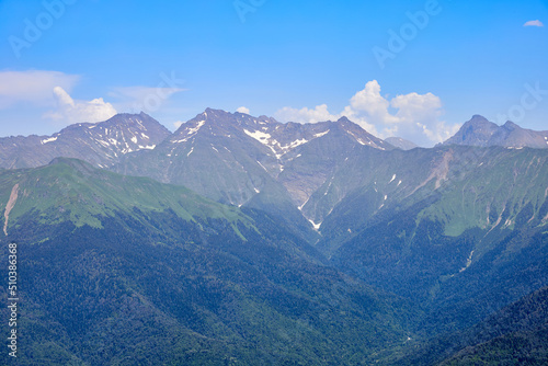 Mountain bright landscape with snow-capped mountains, blue sky and clouds. Mountainside covered with green grass and fir trees.