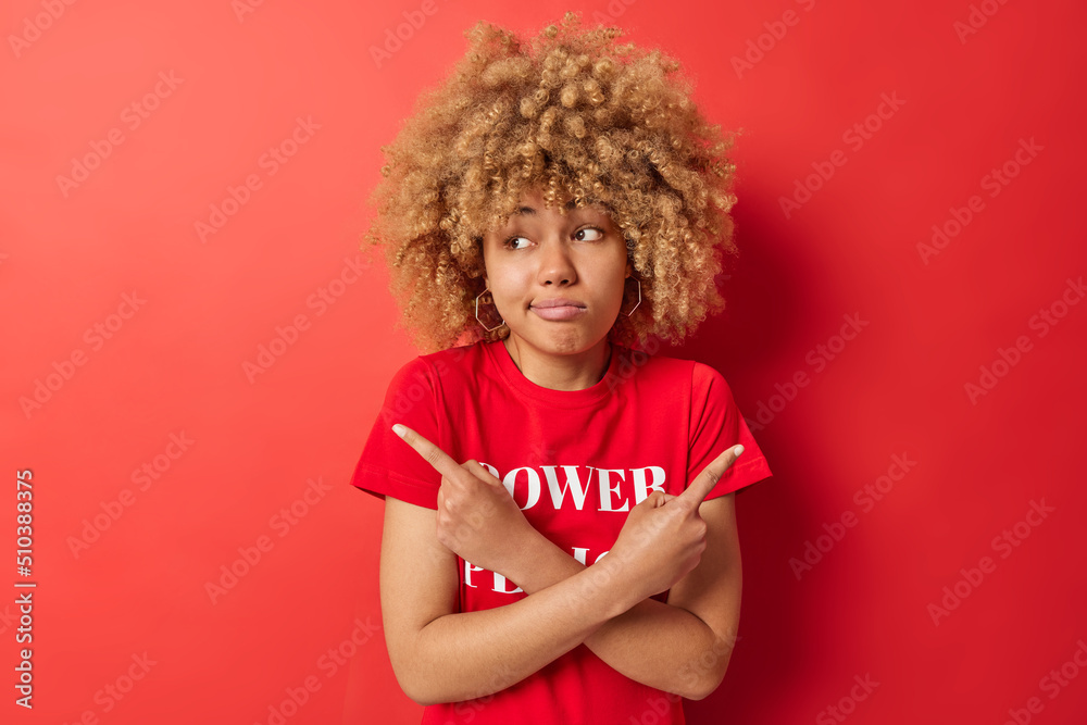 Confused curly haired young European woman points sideways chooses between two options has hesitant expression dressed in casual t shirt isolated over red background. People and choice concept