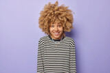 Portrait of glad young European woman with natural curly hair smiles positively dressed in casual striped jumper has good mood isolated over purple background. Human authentic emotions concept