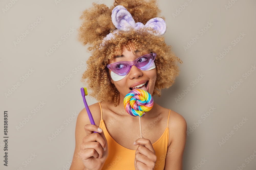 Photo of lovely woman with curly bushy hair wears purple sunglasses headband and orange t shirt licks multicolored lollipop applies beauty patches holds toothbrush isolated over grey background. Photo of lovely woman with curly bushy hair wears purple sunglasses headband and orange t shirt licks multicolored lollipop applies beauty patches holds toothbrush isolated over grey background.