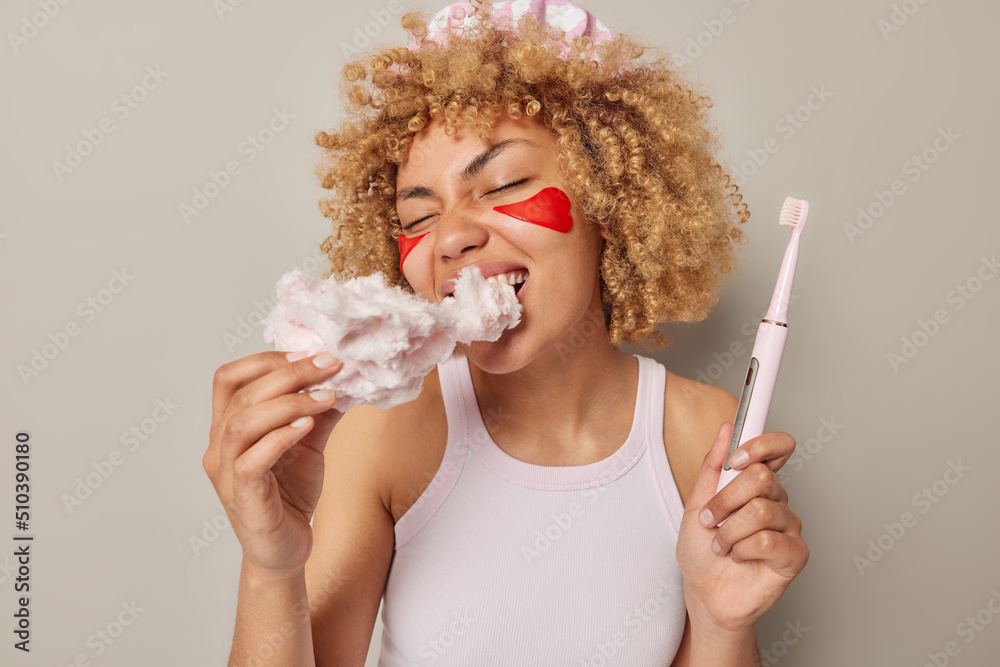Indoor shot of curly haired woman bites sweet candy floss harmful for teeth holds electric brush applies red beauty patches wears casual t shirt isolated over grey background. Unhealthy food - obrazy, fototapety, plakaty 