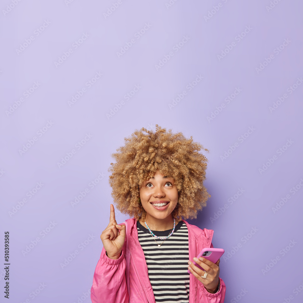 Vertical shot of cheerful young woman dressed in casual clothes uses mobile phone for sending messages points index finger overhead isolated over purple background empty space for your promotion Vertical shot of cheerful young woman dressed in casual clothes uses mobile phone for sending messages points index finger overhead isolated over purple background empty space for your promotion
