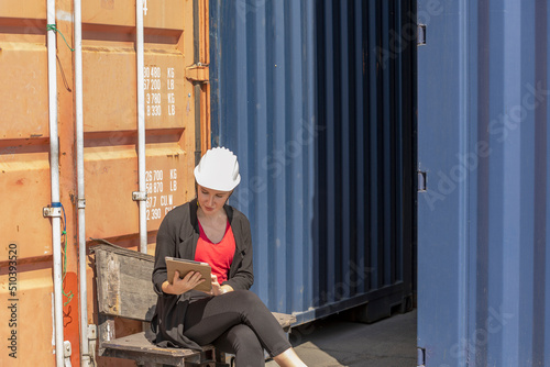 Caucasian female worker uses tablet or smart phone to check stock in a logistic shipyard next to a blue container. Lady in suit with white hard hat uses digital device for her inventory