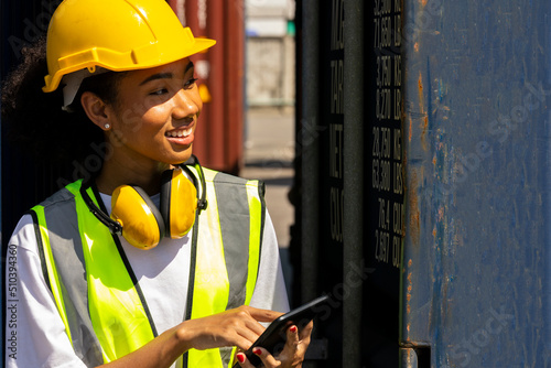 Young African American trainee in safety suit, yellow hard hat and earmuff uses tablet or smart phone in a shipyard. African curly hair female worker in smile. Female loader with yellow earmuff