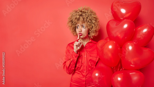 Horizontal shot of curly haired woman has leaked makeup makes silence gesture wears dress holds bunch of heart shaped balloons isolated over red background with empty space for your promotion
