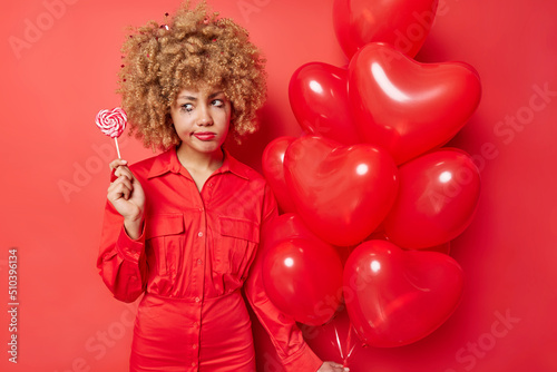 Holiday concept. Sad dissatisfied young woman with curly hair wears dress holds lollipop and bunch of inflated heart balloons isolated over vivid red background feels very tired after party.