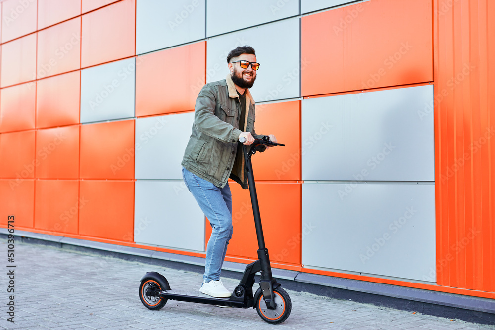 Bearded man in sunglasses posing on electric scooter over orange wall background. Man riding kick scooter. Bearded man in sunglasses posing on electric scooter over orange wall background. Man riding kick scooter.
