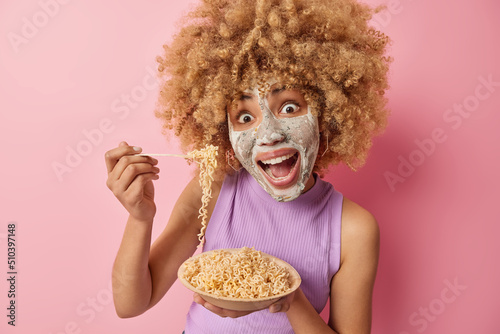Amazed woman has curly bushy hair eats delicious noodles exclaims loudly applies beauty mask on face for reducing fine lines dressed in casual t shirt isolated on pink background. Skin care procedures