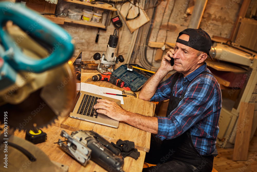 Senior carpenter works on the computer and talks phone with client in the workshop Senior carpenter works on the computer and talks phone with client in the workshop