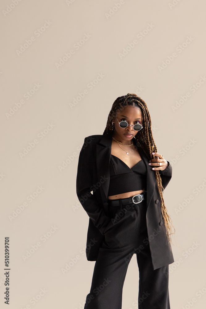 African girl in a black suit with long hair and sunglasses stands on a beige background African girl in a black suit with long hair and sunglasses stands on a beige background