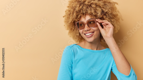 Horizontal shot of cheerful curly haired young European woman wears sunglasses and casual blue jumper concentrated away isolated over beige background empty space for your promotional content