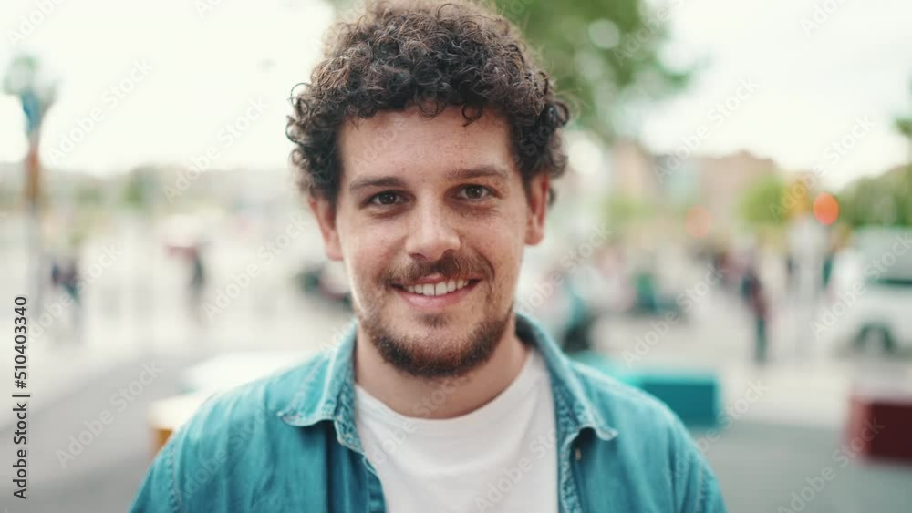 Frontal closeup portrait of young bearded man in denim shirt posing and smiling at camera on modern cityscape background.