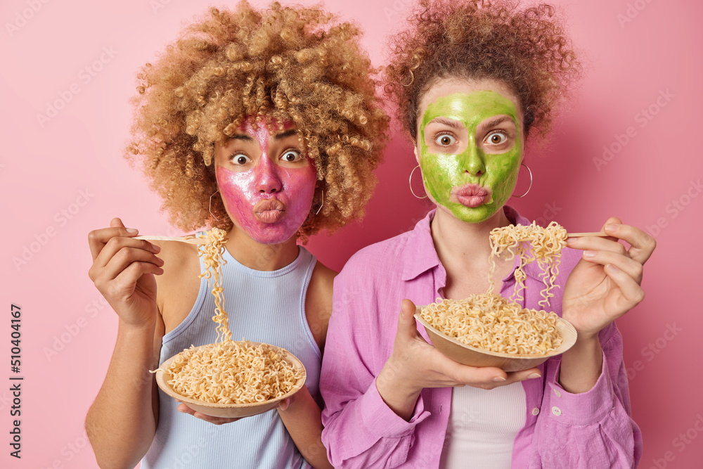 Photo of amazed women with curly hair apply facial masks keep lips folded eat pasta for supper dressed in casual wear take care of skin isolated over pink background. Beauty treatments concept