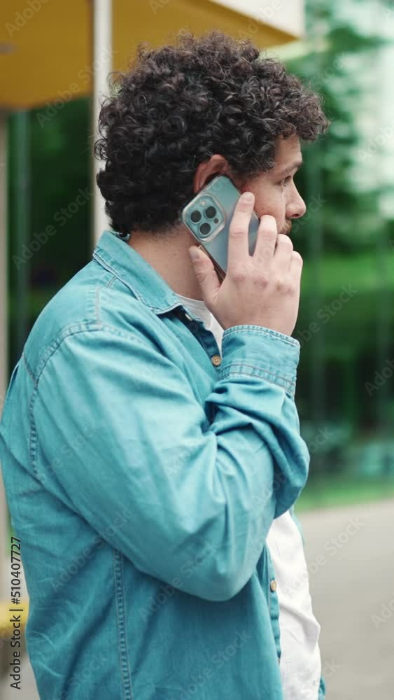 VERTICAL VIDEO: Close-up, young bearded man in denim shirt stands at bus stop waiting for bus and talking on mobile phone on modern city background VERTICAL VIDEO: Close-up, young bearded man in denim shirt stands at bus stop waiting for bus and talking on mobile phone on modern city background