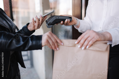 Closeup of female standing outdoors by cafe and paying with smartphone. Cashier hand holding credit card reader machine while client holding phone for NFC payment. Focus on hand with package