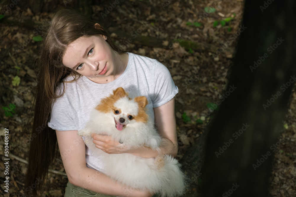 Young woman with long straight hair and fluffy small dog for walk in forest. Pomeranian with his owner for walk. Satisfied dog smiles and sticks out his tongue. Woman and dog looks at camera Young woman with long straight hair and fluffy small dog for walk in forest. Pomeranian with his owner for walk. Satisfied dog smiles and sticks out his tongue. Woman and dog looks at camera