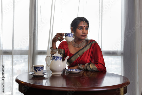 Indian woman wearing red orange saree jewellery choker set necklace jhumka earring maang tikka waist chain stand pose look see smile mood expression siting vintage table tea pot cup bright window back photo