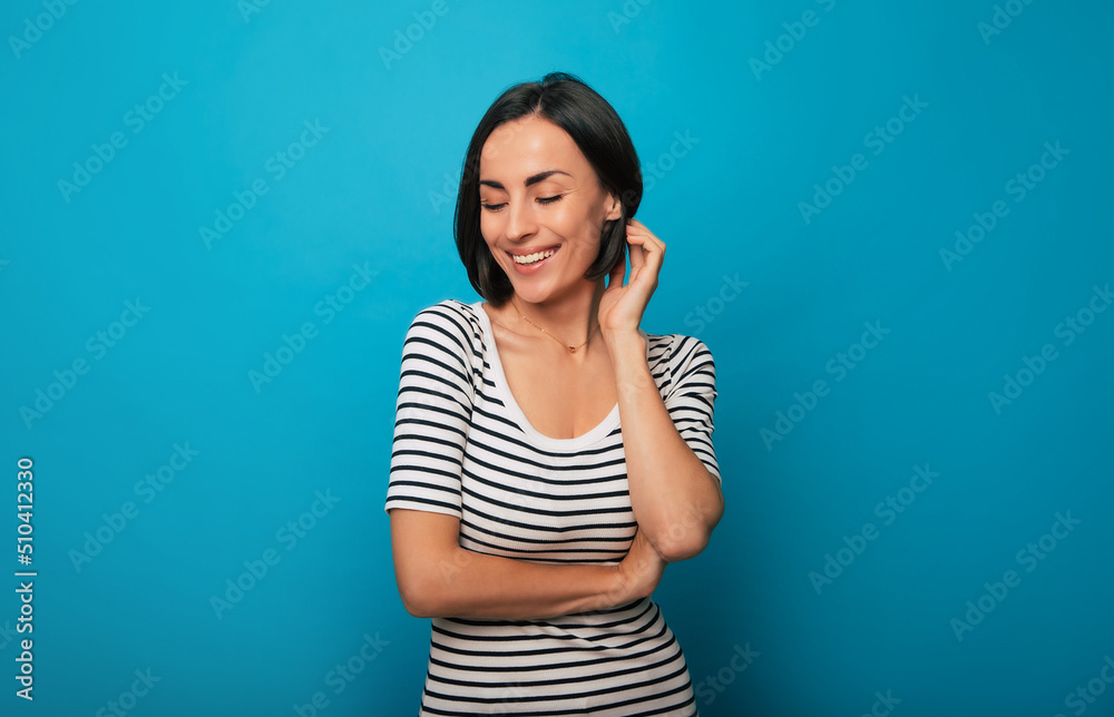 Close up portrait of a gorgeous smiling young brunette woman while she is posing in a striped t-shirt with crossed arms and isolated on blue background