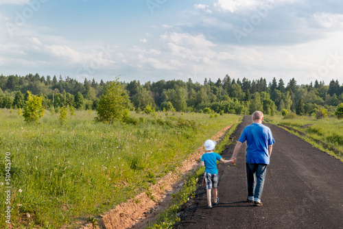 rear view of a father and son walking along the road and holding hands in a forest strip