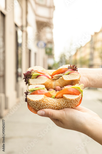 Bagel with prosciutto, cream cheese in woman hand. Holding a Sandwich with lettuce for breakfast on street