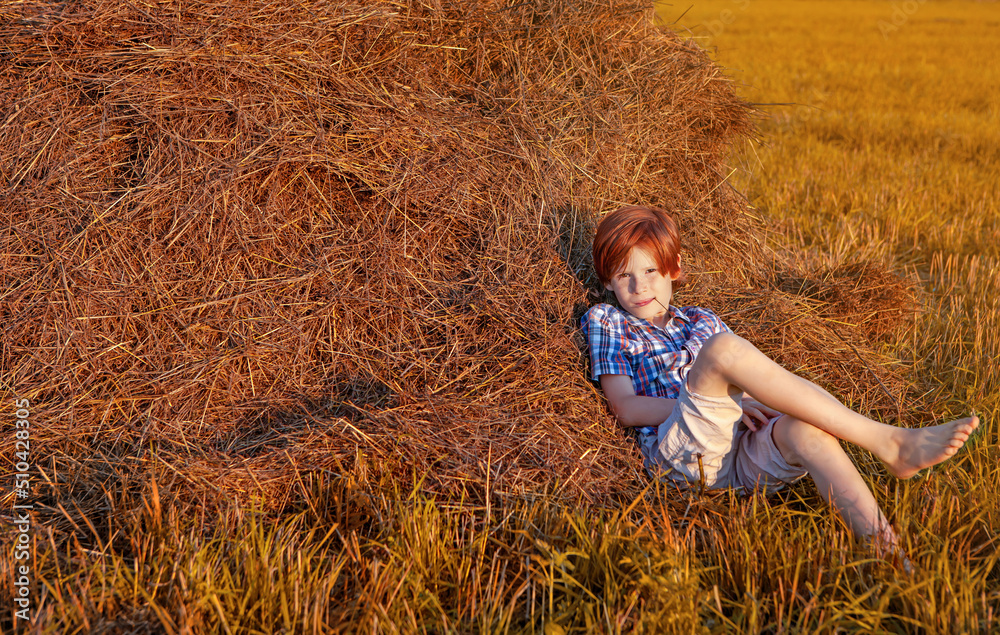 A red-haired boy leaning against a haystack on a lazy summer evening at sunset A red-haired boy leaning against a haystack on a lazy summer evening at sunset