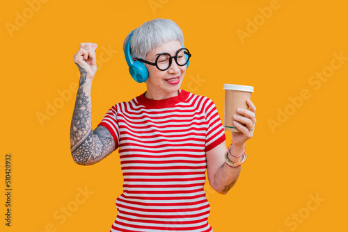 Cheerful elderly gray-haired asian female woman with tattoo casual cloth listening music with headphones keeping eyes closed isolated on pastel yellow color background studio portrait