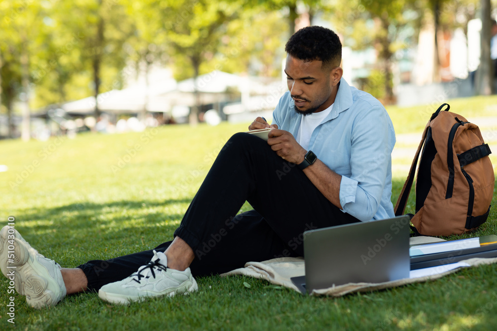 African american male student writing something in notebook, sitting with laptop in park and studying outdoors African american male student writing something in notebook, sitting with laptop in park and studying outdoors