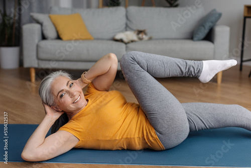 Active senior woman doing abs exercises on yoga mat at home and smiling at camera, working out in living room