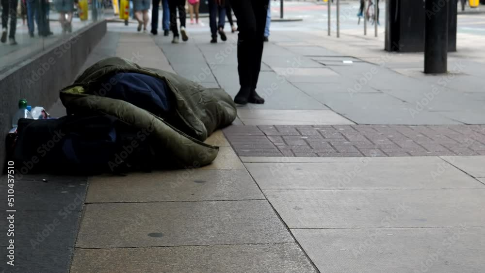 Legs of people walking past homeless person sleeping rough on pavement by store front window in Manchester city downtown.