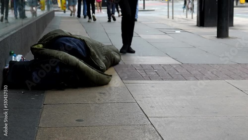 Legs of people walking past homeless person sleeping rough on pavement by store front window in Manchester city downtown.
