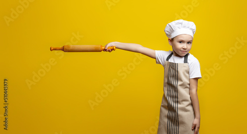 Funny 6-7-year-old Caucasian little chef in uniform holding a rolling pin pointing to the side, isolated on a yellow background. concept of favorite food for children. Copy space photo