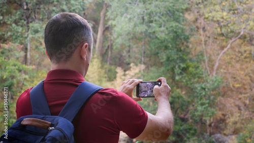 Hiker blogger or vlogger man in woods shooting forest. Male traveler videographer with backpack, standing in nature and filming video on smartphone camera. Back view