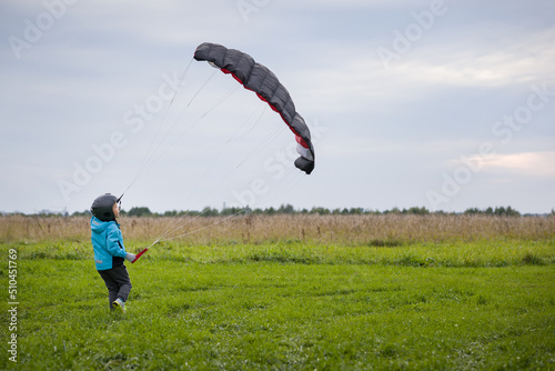 A cute kid in a blue jacket runs across a green field, launches a training parachute wing, holds the brakes, catches the wind and controls the kite, enjoying the flight in the air over the open area