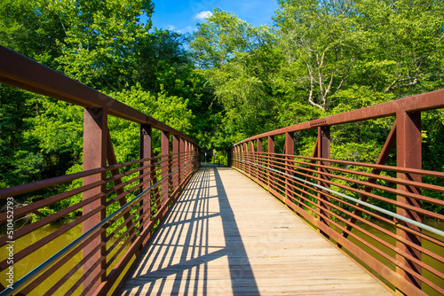 a long metal rust colored bridge over the silky brown waters of Little River surrounded by lush green trees, grass and plants with blue sky and clouds at Olde Rope Mill Park in Woodstock Georgia  photo